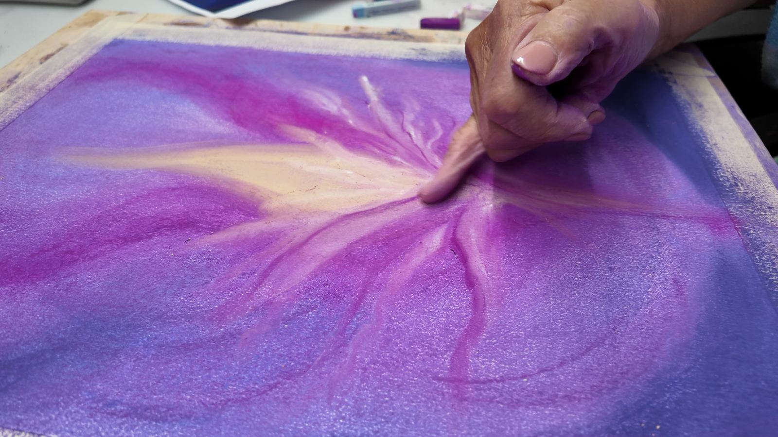 Des doigts estompent du pastel violet et doré sur une feuille, pendant un atelier